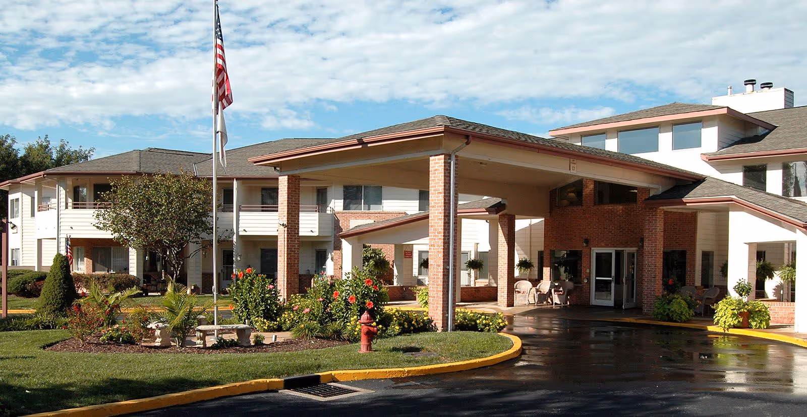 Front entrance of a senior living building with a covered porte-cochere, flagpole, and landscaped gardens.