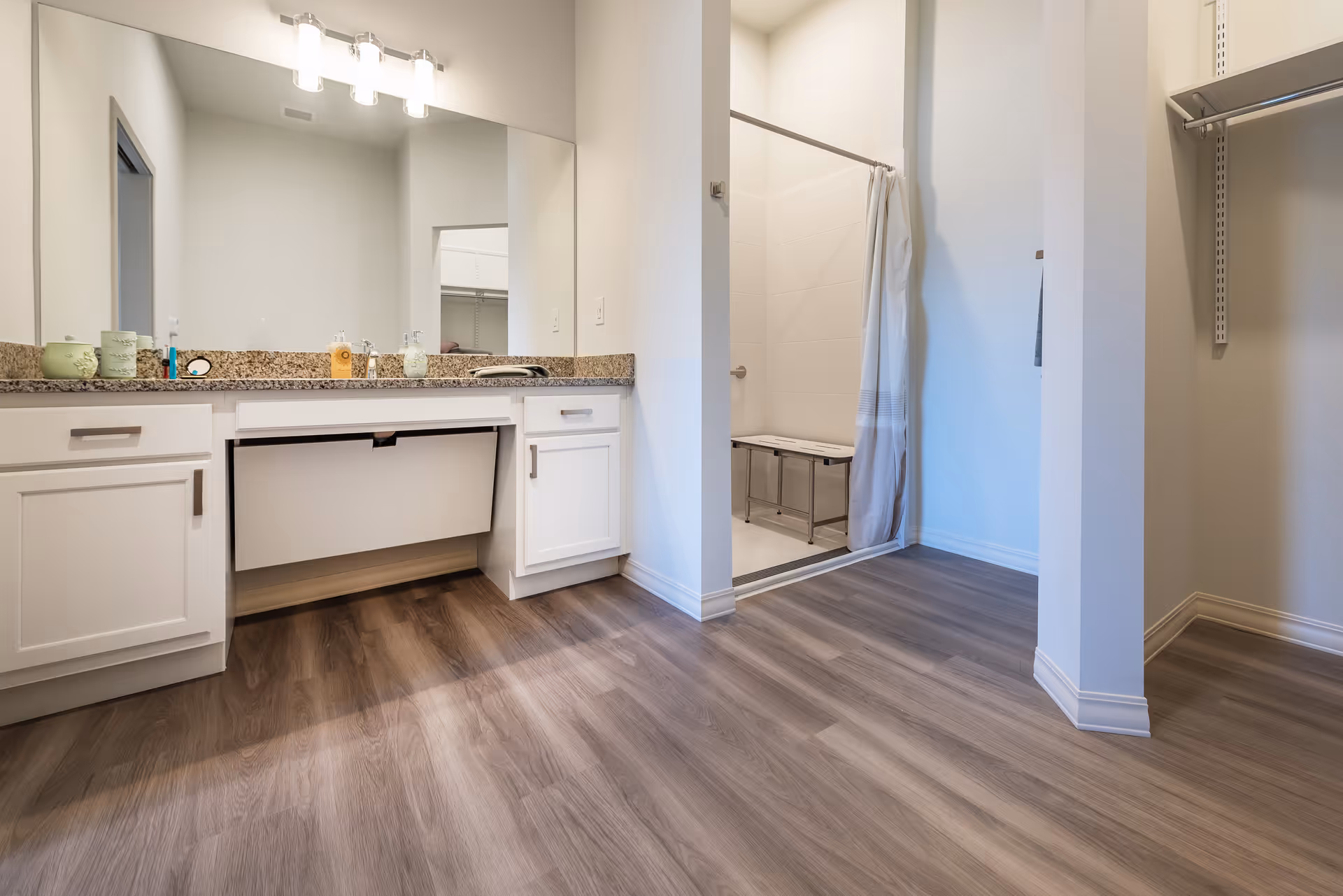 A clean and modern bathroom with a large mirror above a granite countertop with white cabinets. There is a walk-in shower with a white curtain and a metal bench inside. The floor is covered with wood-like vinyl planks, and there is an open closet area with shelving on the right side.