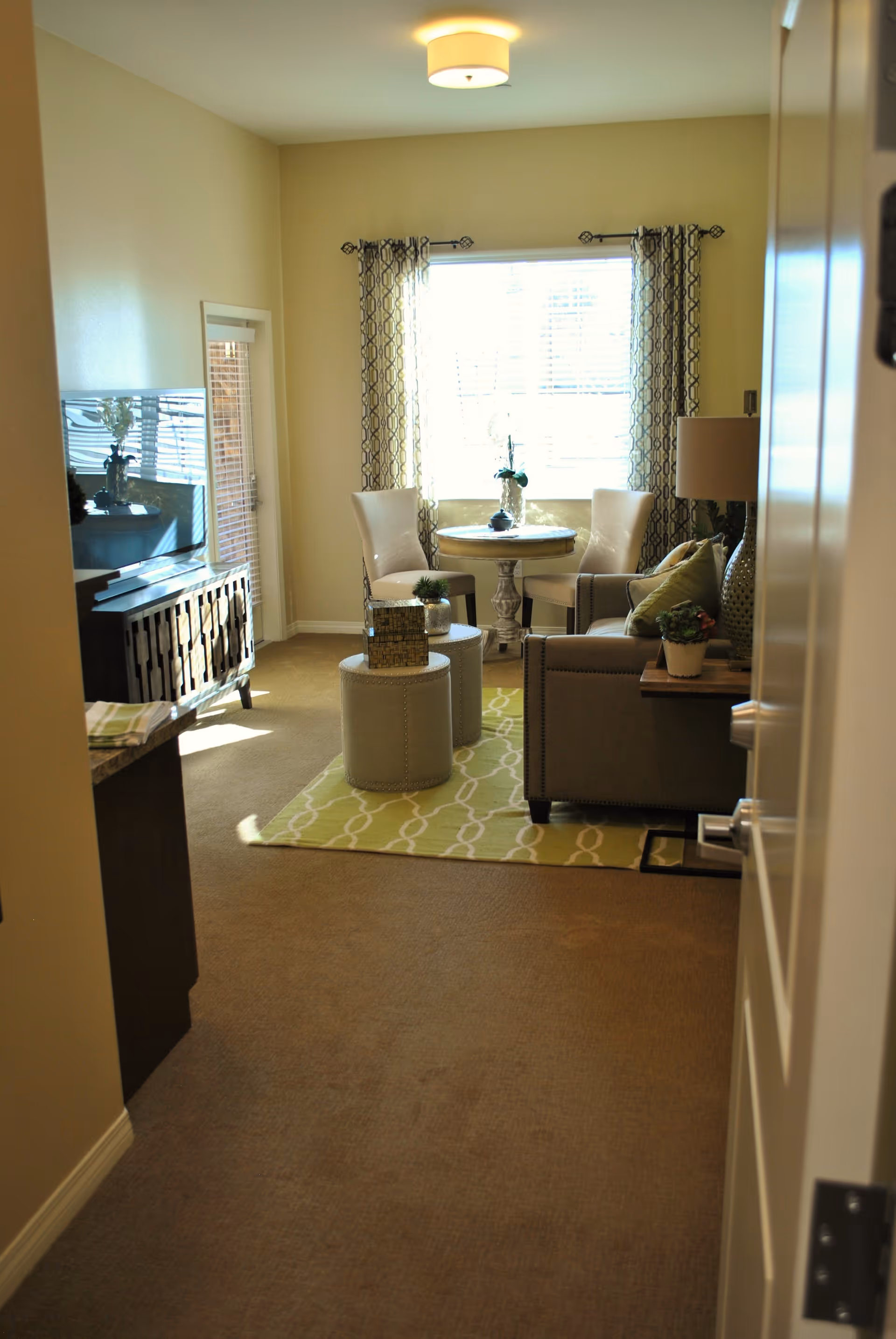 Interior view of a living room area with beige walls and carpet. The room features a small round table with two white chairs near a window with patterned curtains. There is a gray armchair with green pillows, a lamp on a side table, two round ottomans on a green patterned rug, and a TV on a wooden stand to the left.