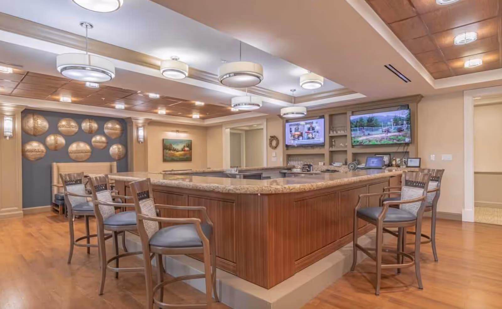 A spacious interior room with a large wooden counter in the center surrounded by high chairs. The room features modern ceiling lights, wood flooring, and decorative wall art with circular metallic pieces. Two flat-screen TVs are mounted on the wall behind the counter, displaying different images.