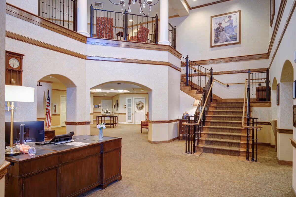 Interior view of a senior living facility lobby with a wooden reception desk, a computer, a lamp, and a vase with flowers. There is a staircase with a carpet runner leading to an upper floor with a railing. The walls are decorated with framed artwork and a clock. The area is well-lit with ceiling lights and a chandelier.