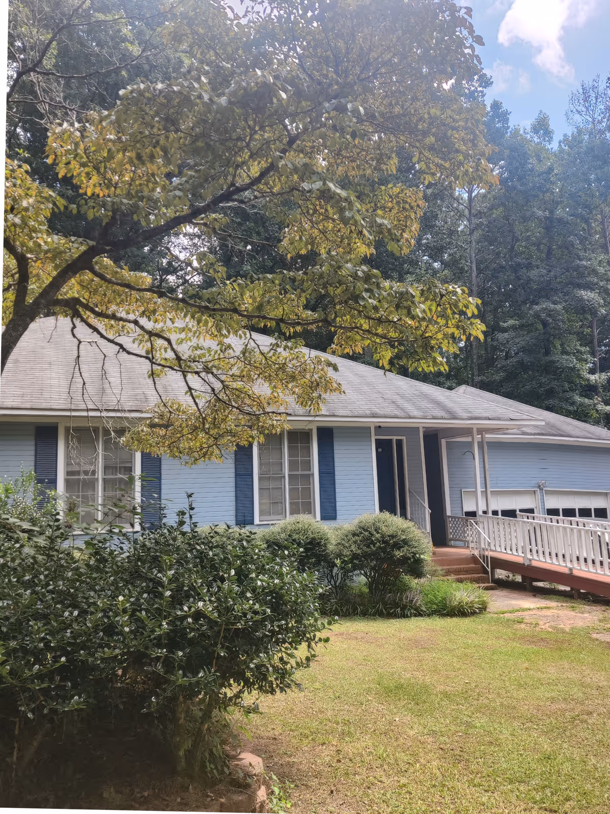 Single-story light blue house with a front porch, wheelchair ramp, garage and surrounding trees and shrubs.