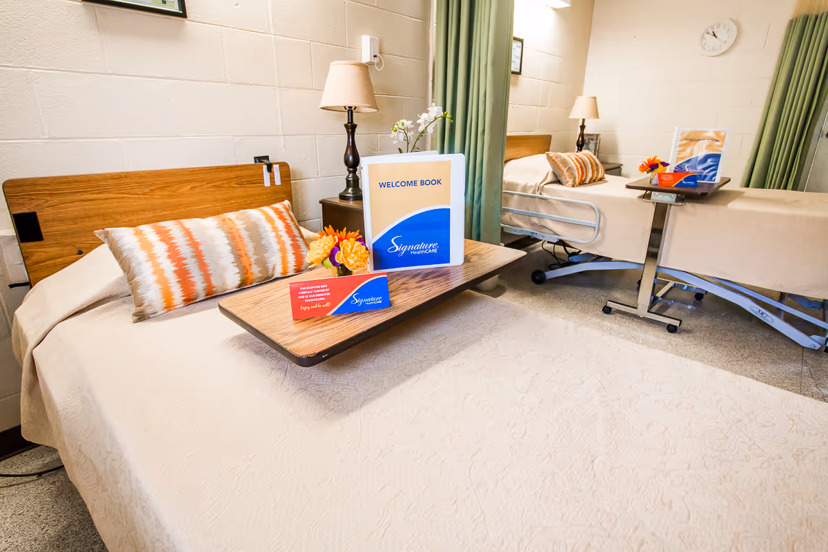 A bright and clean room with two hospital-style beds, each with a wooden headboard, beige bedspreads, and decorative pillows. Each bed has a wooden overbed table holding a welcome book, a small flower arrangement, and a red card. There are two bedside tables with lamps, green curtains, and a wall clock in the background.