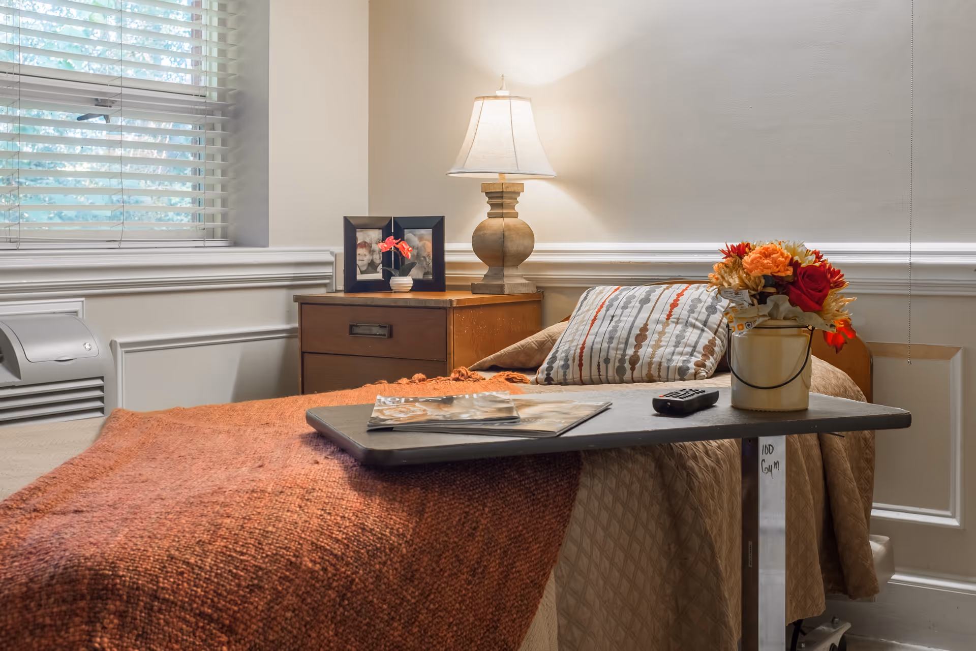 A cozy bedroom with a single bed covered in a brown quilt and an orange blanket. A bedside table holds a lamp, two framed photos, and a small potted plant. A tray table on the bed has a remote control, a magazine, and a vase with colorful flowers. A window with white blinds is partially visible on the left side.