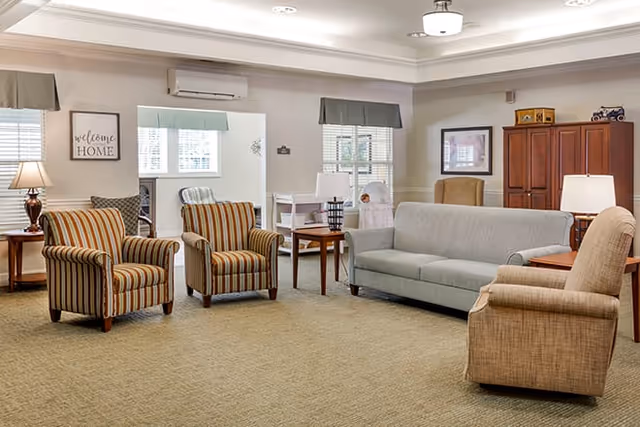 A cozy living room with a beige carpet, a gray sofa, two striped armchairs, and a beige armchair. There are side tables with lamps, a wooden cabinet, and windows with blinds and valances. A wall sign reads 'welcome HOME'.