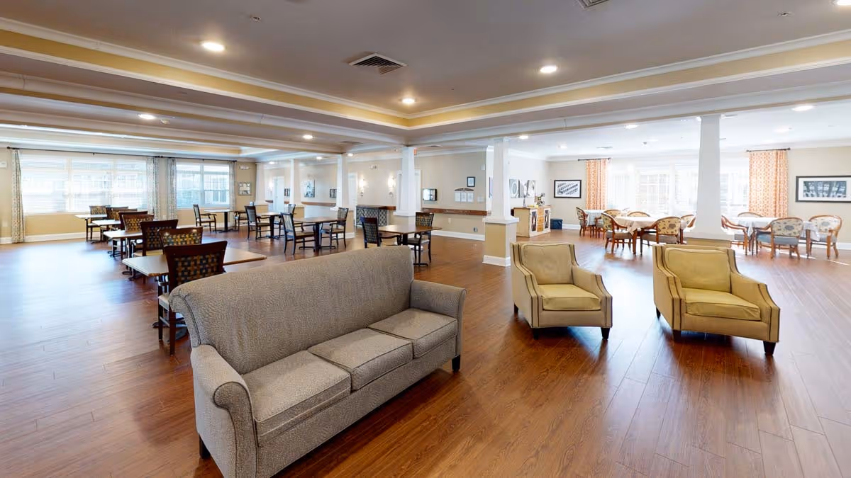 A spacious and well-lit common area in a senior living facility featuring a gray sofa and two beige armchairs in the foreground. In the background, there are multiple dining tables with chairs arranged neatly on a wooden floor, with large windows allowing natural light to fill the room. The ceiling has recessed lighting and decorative molding.