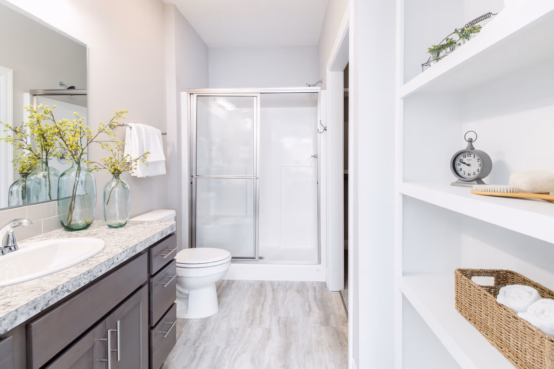 Bright modern bathroom with a vanity and sink, toilet, glass-enclosed shower, and open shelving with decor.
