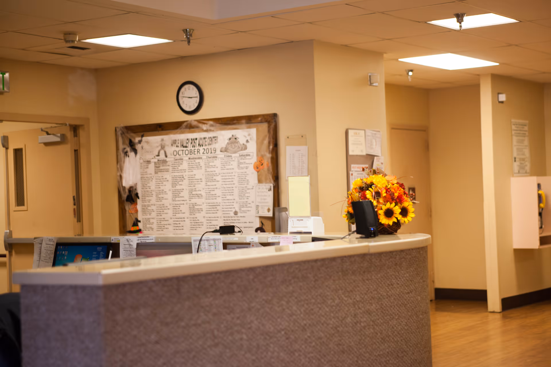 Reception desk area in a care facility with a bulletin board, wall clock, computer monitors, and a bouquet of sunflowers on the counter.