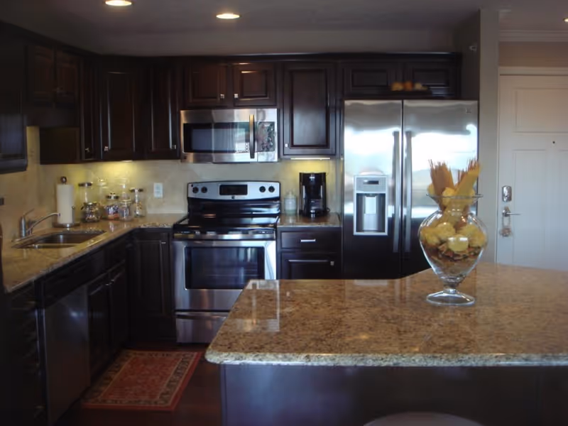 Modern kitchen with dark wood cabinets, stainless steel appliances including a refrigerator, stove, microwave, and dishwasher. There is a granite countertop island with a glass vase holding decorative items. The kitchen has a sink with a paper towel holder and jars on the counter.