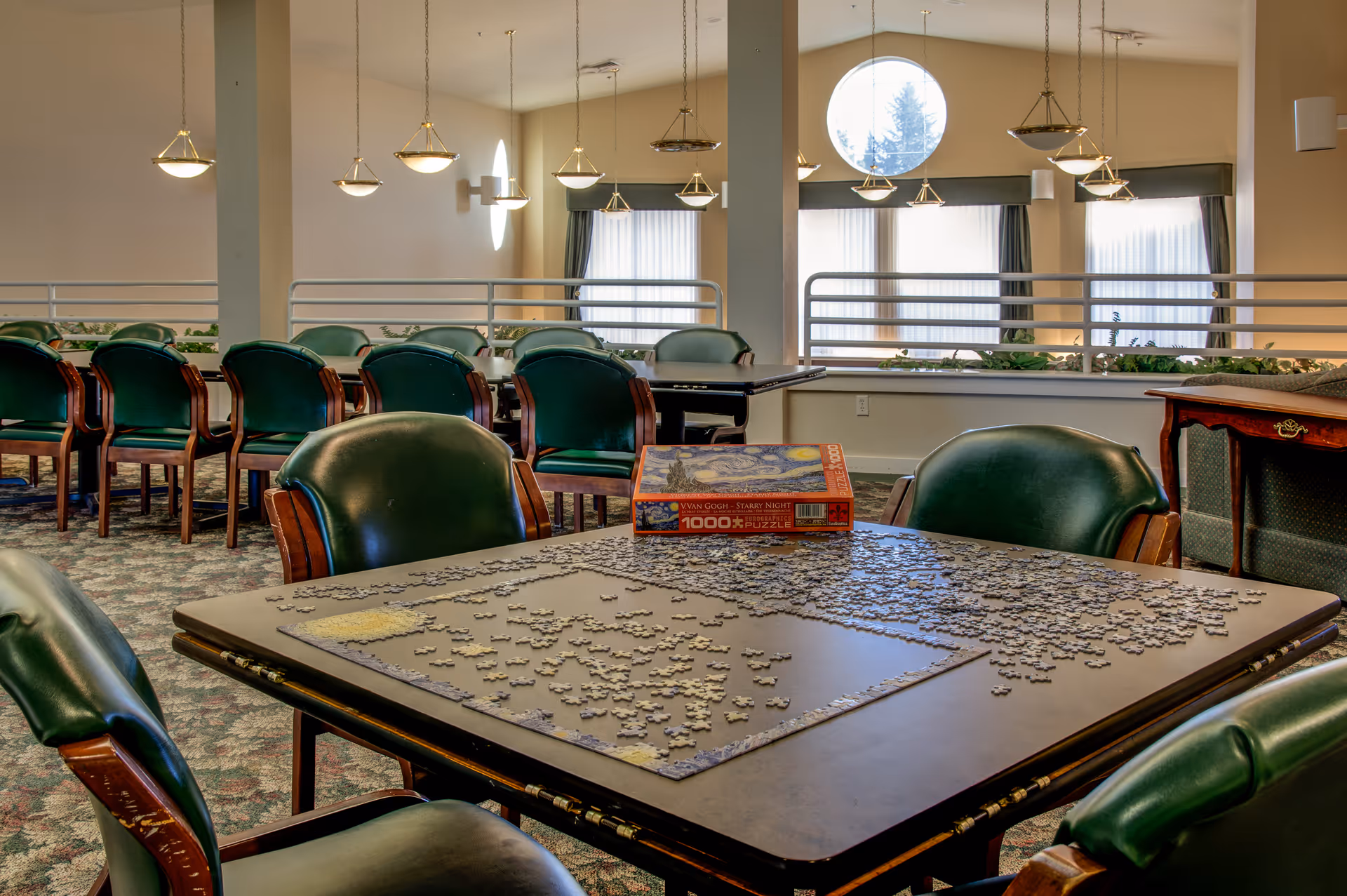 A bright common room with green upholstered chairs and a table in the foreground covered with jigsaw puzzle pieces and its box.