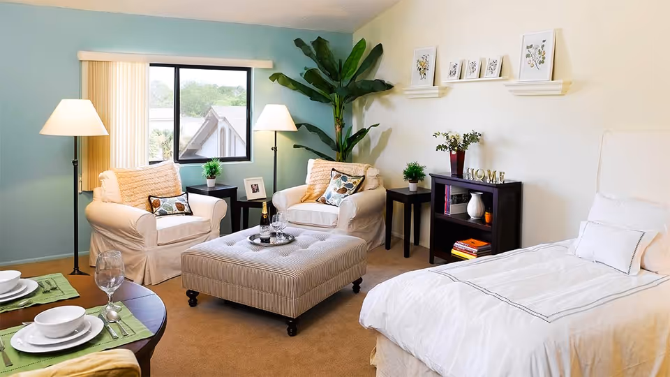 A cozy and well-lit senior living room and bedroom area featuring a single bed with white bedding, two cushioned armchairs with decorative pillows, a large ottoman in the center, a small dining table set for two, floor lamps, potted plants, framed botanical artwork on the wall, and a window with vertical blinds showing an outdoor view.