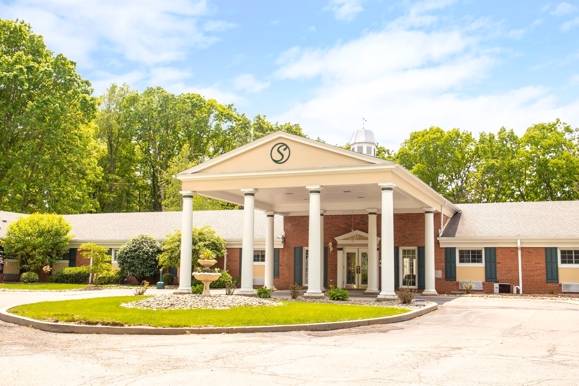 Front exterior view of Shelbourne Personal Care facility showing a single-story brick building with a covered entrance supported by white columns, surrounded by green trees and a circular driveway with a small garden and a decorative fountain.