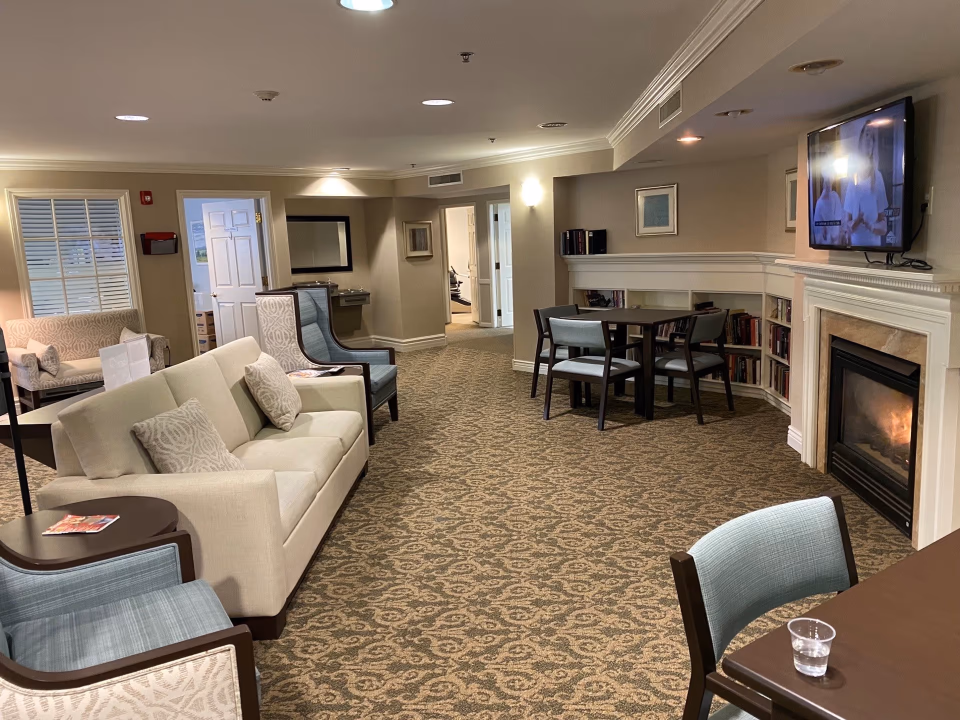 A cozy senior living facility common area with beige walls and patterned carpet. The room features a beige sofa with patterned pillows, several upholstered chairs in light blue and beige, a small round table with chairs near a fireplace, and a wall-mounted TV above the fireplace. There are bookshelves built into the wall under the TV and fireplace. The room is well-lit with ceiling lights and wall sconces, and there are doorways leading to other rooms in the background.