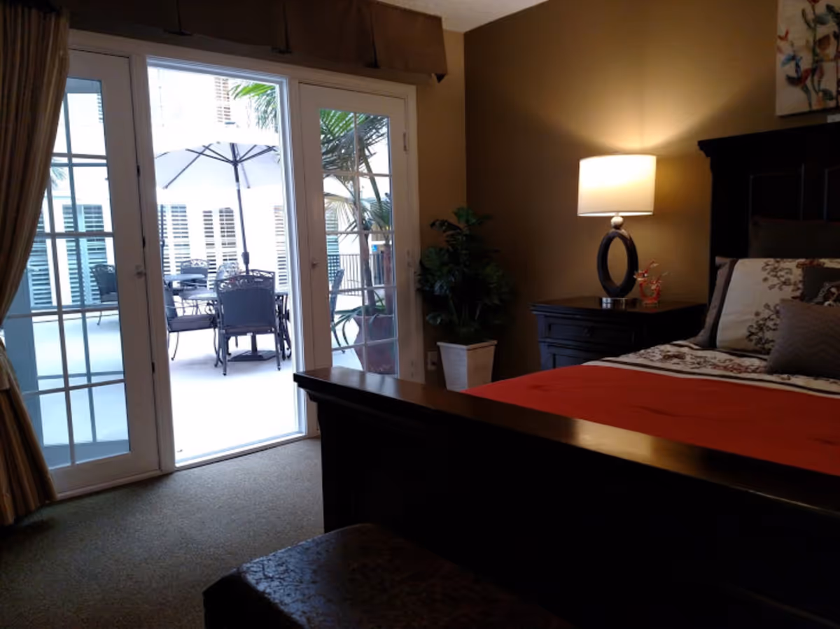 A bedroom with a dark wooden bed frame and a red bedspread. There is a nightstand with a modern lamp and decorative items next to the bed. A potted plant is placed near the sliding glass doors that lead to an outdoor patio with a table, chairs, and an umbrella.