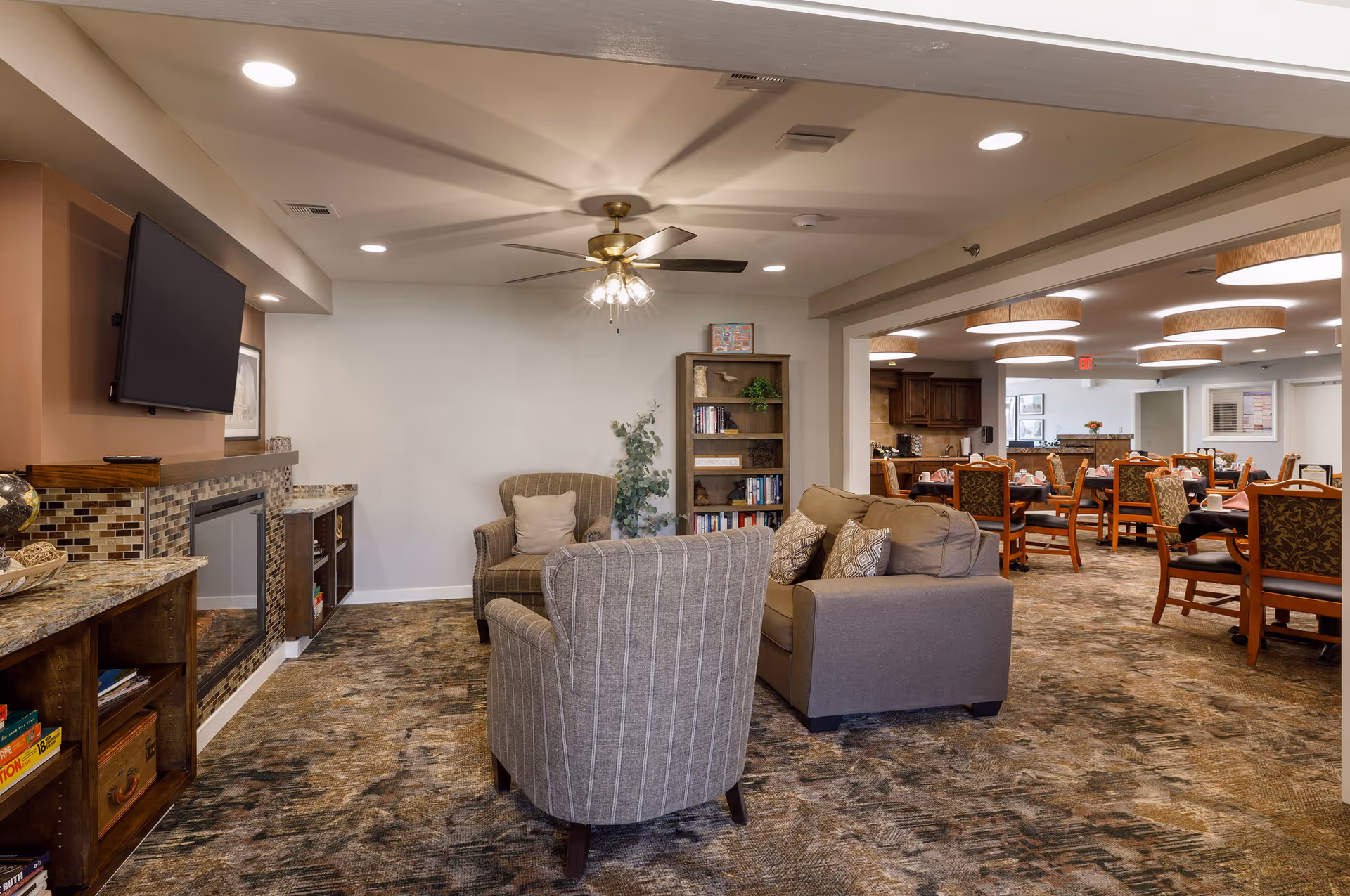 A cozy living room area with a striped armchair, a cushioned sofa with patterned pillows, a bookshelf, and a ceiling fan with lights. Adjacent to the living room is a dining area with multiple tables and chairs, illuminated by round ceiling lights. The living room also features a wall-mounted TV above a fireplace with a mosaic tile surround.