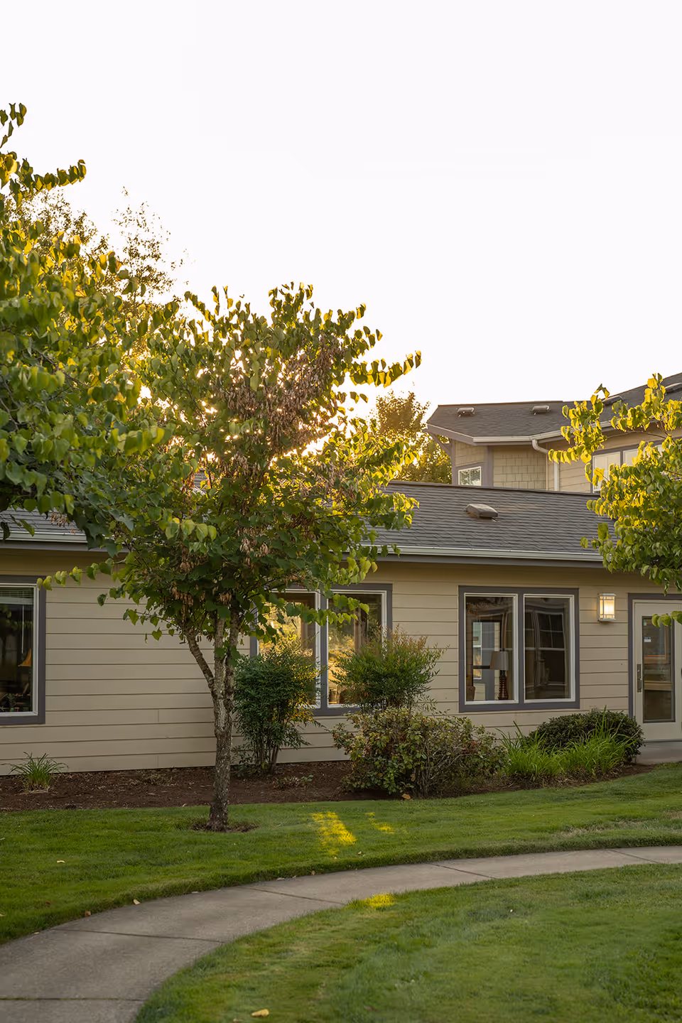 Front exterior of a single-story assisted living building with windows, shrubs, trees, and a curved sidewalk.