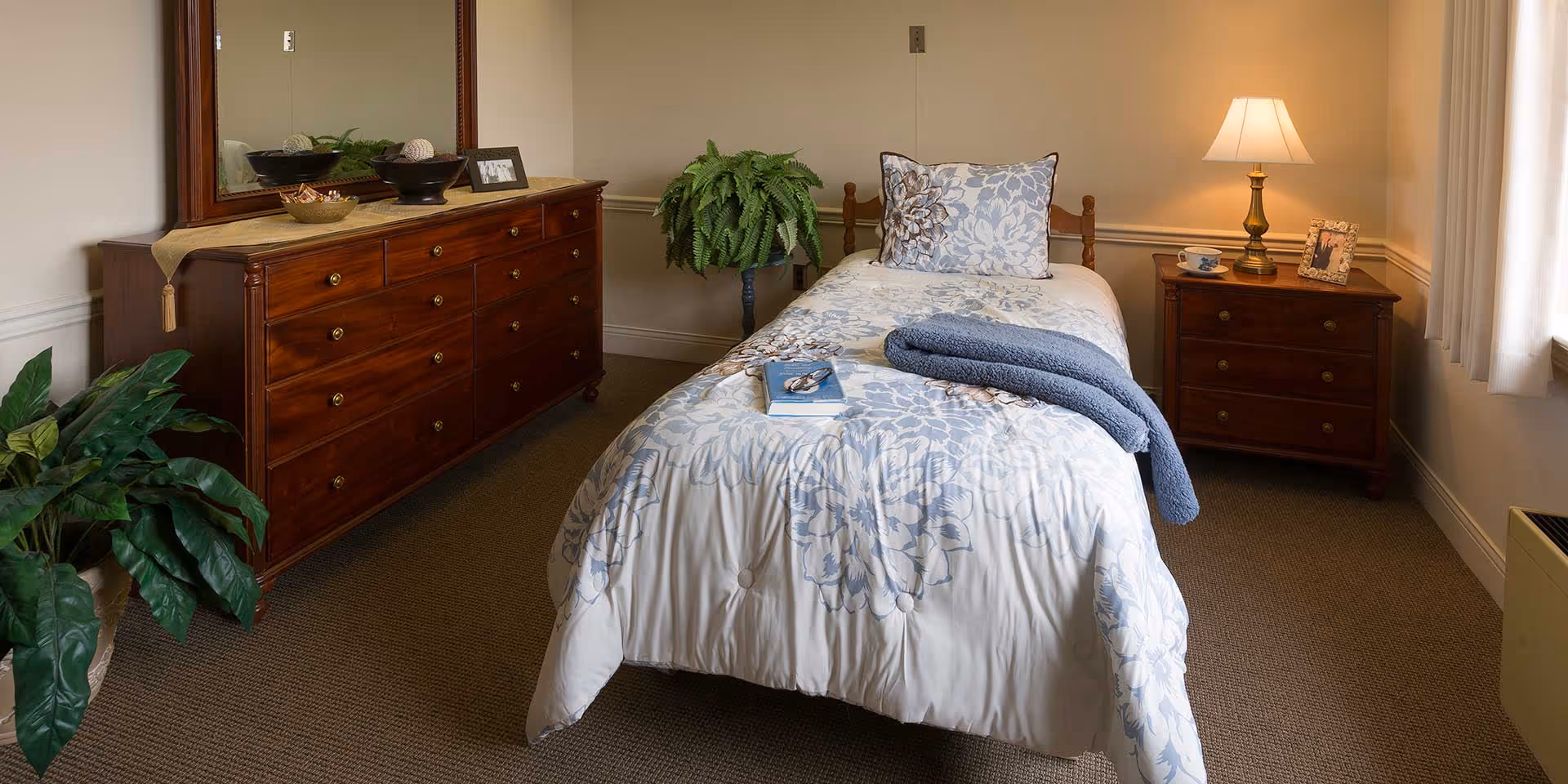 A cozy bedroom with a single bed covered in a floral patterned comforter and a matching pillow. A folded blue blanket and a book rest on the bed. To the right of the bed is a wooden nightstand with a lamp, a teacup, and a framed photo. On the left side of the room is a wooden dresser with a large mirror above it, decorated with bowls and a small framed photo. There are two green plants in the room, one on the floor and one on a stand. The room has beige walls and carpeted floor, with a window covered by white curtains on the right.