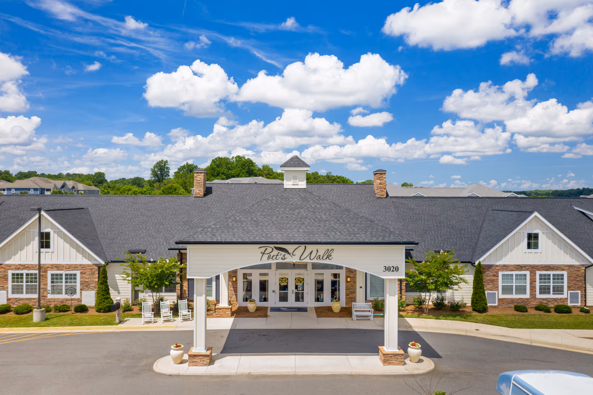 Front exterior view of Poet's Walk Warrenton, A Memory Care Community, showing a single-story building with a covered entrance, white pillars, and a sign with the facility name. The building has a dark shingled roof, brick and white siding, and is surrounded by small trees and shrubs under a blue sky with scattered clouds.