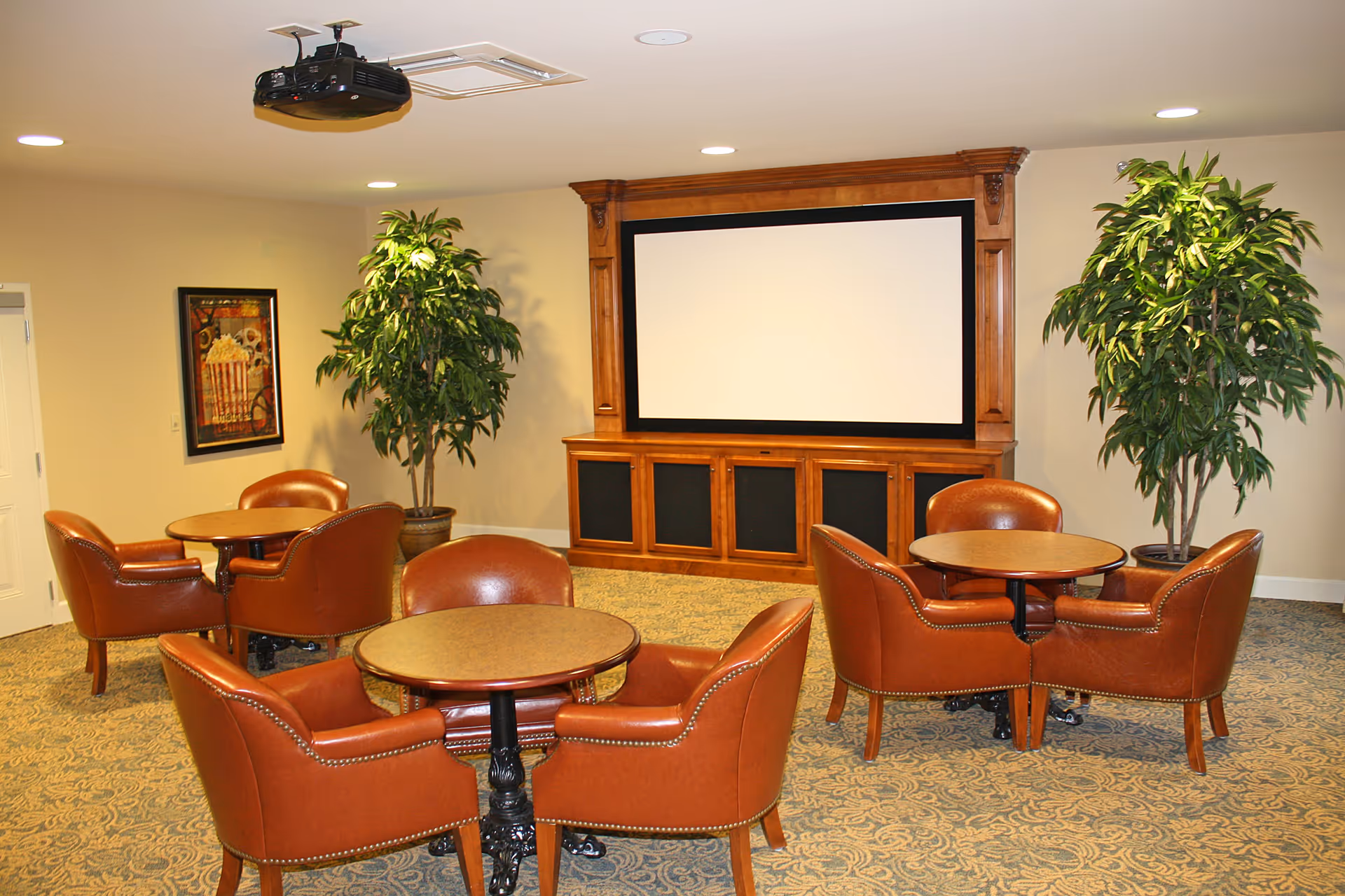 A cozy common room with three round tables, each surrounded by four brown leather chairs. There is a large wooden entertainment center with a blank projection screen on the wall. Two large potted plants flank the entertainment center, and a projector is mounted on the ceiling. The room has patterned carpet and recessed lighting.
