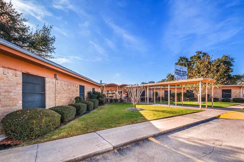 Exterior view of a single-story brick building with a covered walkway, neatly trimmed bushes, and a small lawn area under a clear blue sky.