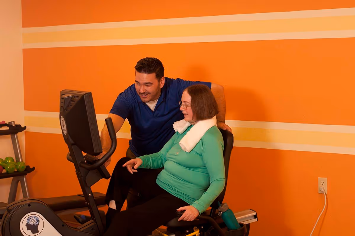 A man in a blue shirt assists a woman in a green long-sleeve shirt with a white towel around her neck as she uses a recumbent exercise bike in a room with orange and yellow striped walls.