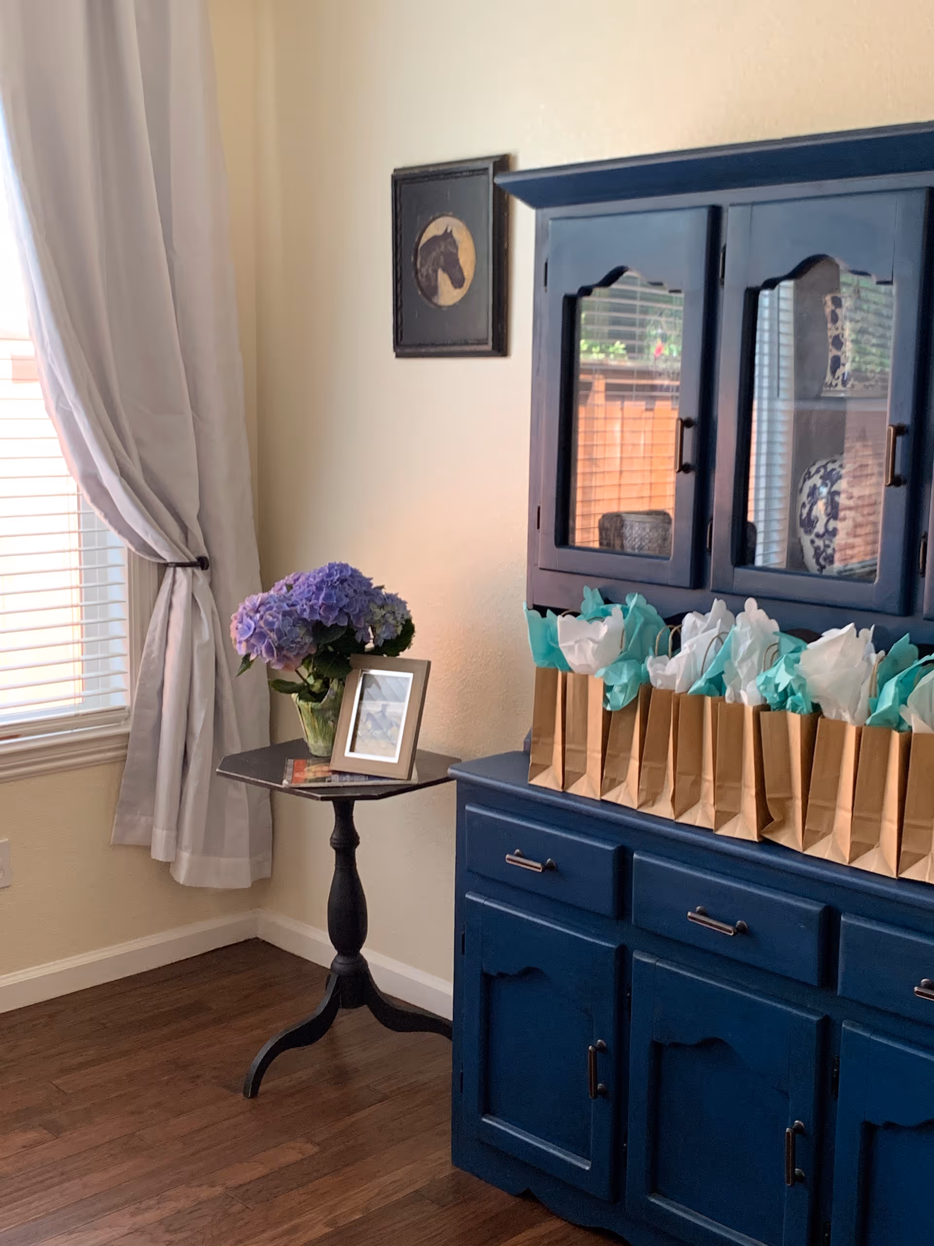 A cozy corner of a room featuring a dark blue wooden cabinet with glass doors, topped with several brown paper gift bags filled with white and teal tissue paper. Next to the cabinet is a small round black table holding a vase of purple hydrangeas and a framed photo. A window with white blinds and light gray curtains is visible on the left, and a framed horse silhouette hangs on the beige wall above the cabinet.