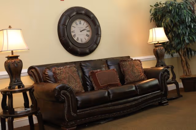 Leather sofa with decorative pillows flanked by table lamps, a large wall clock, and a potted plant in a sitting area.