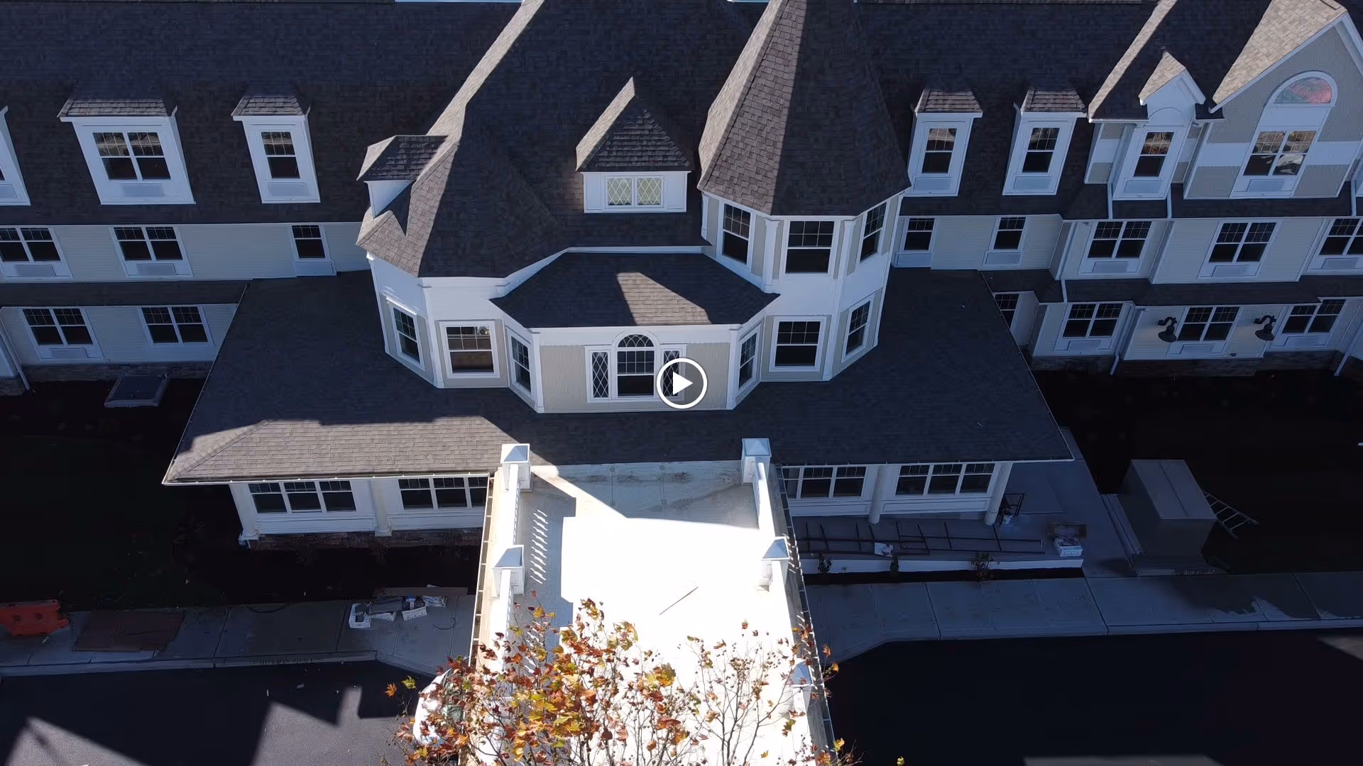 Aerial view of a large, multi-story residential building with a complex roof design, multiple windows, and a driveway leading to the entrance. The building appears to be a senior living facility.