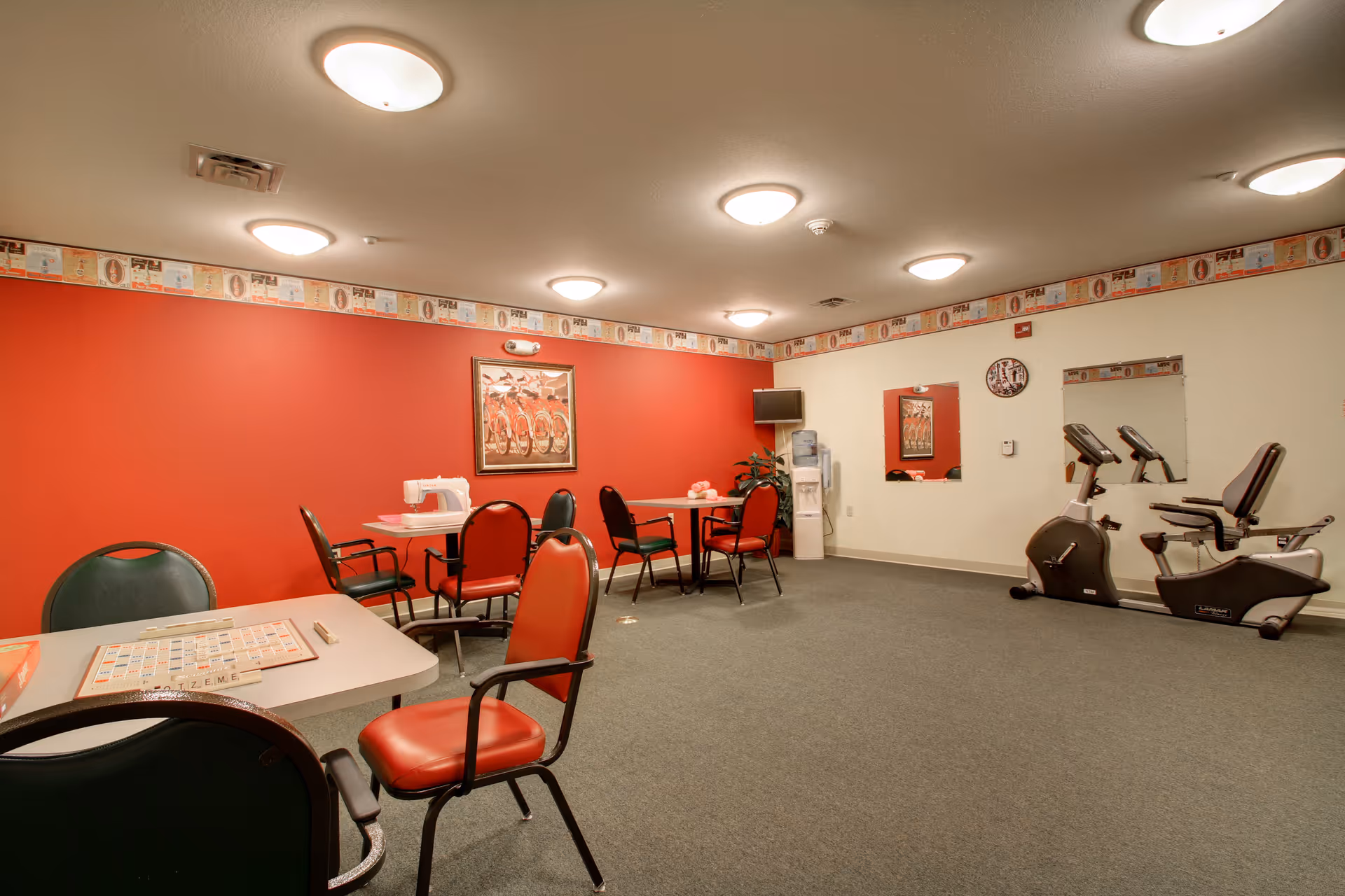 A senior living community room with tables and chairs set for games, exercise bikes, and a water cooler against a red accent wall.