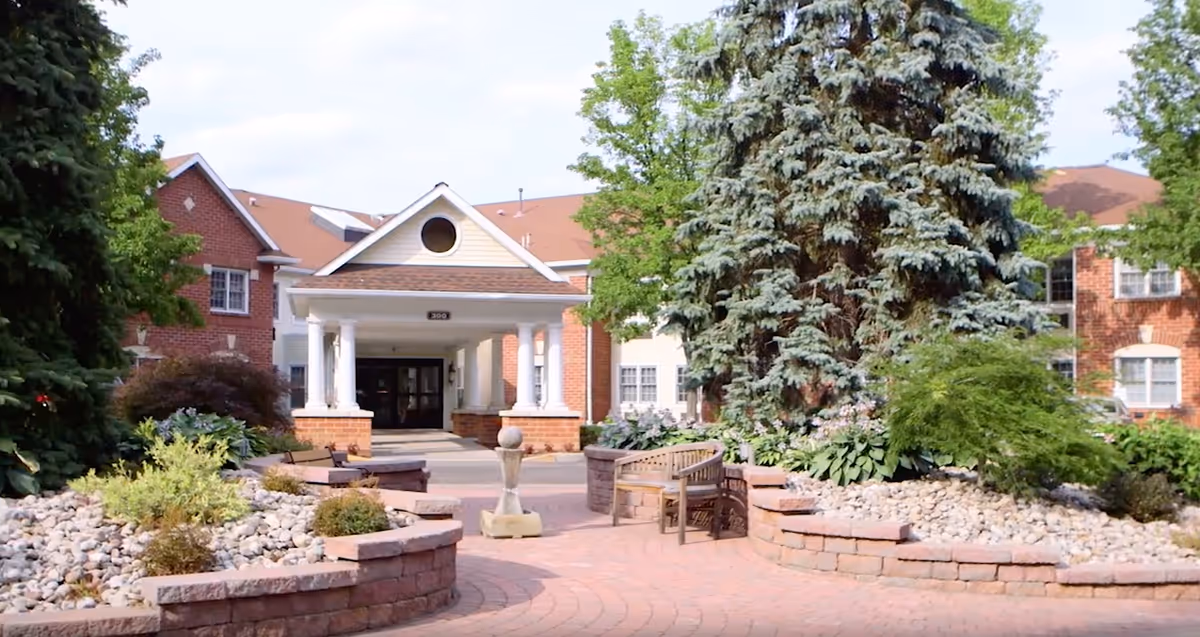 Front entrance of a brick senior living building with a covered portico, landscaped stone planters, benches, and a large evergreen tree.