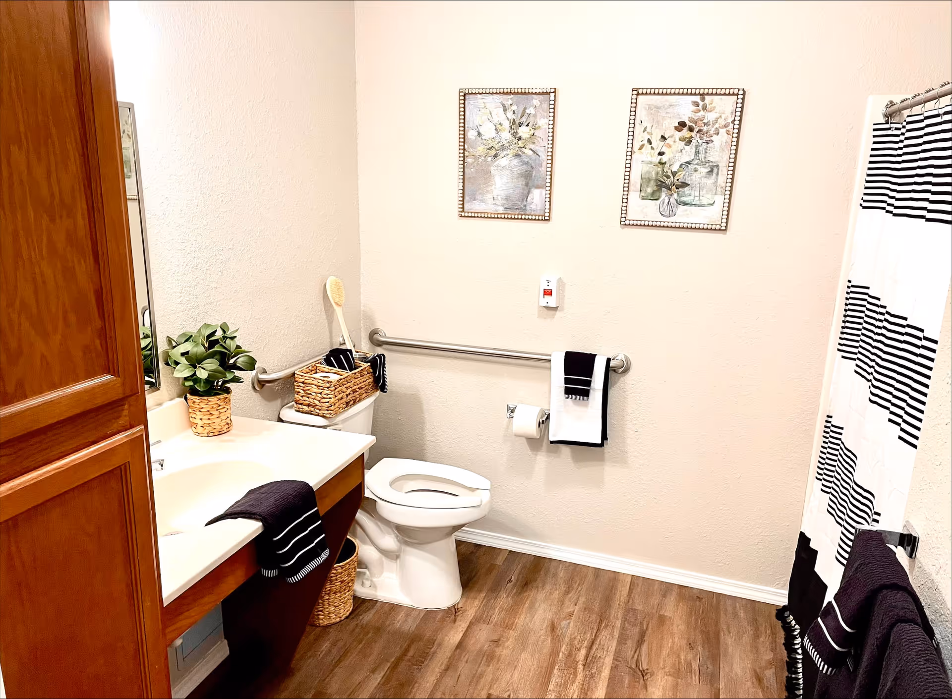 A clean and well-lit bathroom with a white toilet, a wooden vanity with a white countertop, a sink, and a mirror. There are two framed floral pictures on the wall above a grab bar with a towel and toilet paper holder. A black and white striped shower curtain is visible on the right side. The floor has wood-like vinyl flooring, and there are decorative baskets and a plant on the vanity and toilet tank.