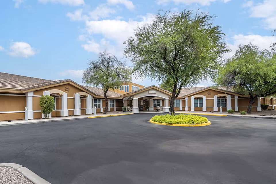 Exterior view of Brookdale Tempe senior living facility showing a single-story building with a covered entrance, surrounded by trees and a circular driveway with a landscaped island in the center under a partly cloudy sky.