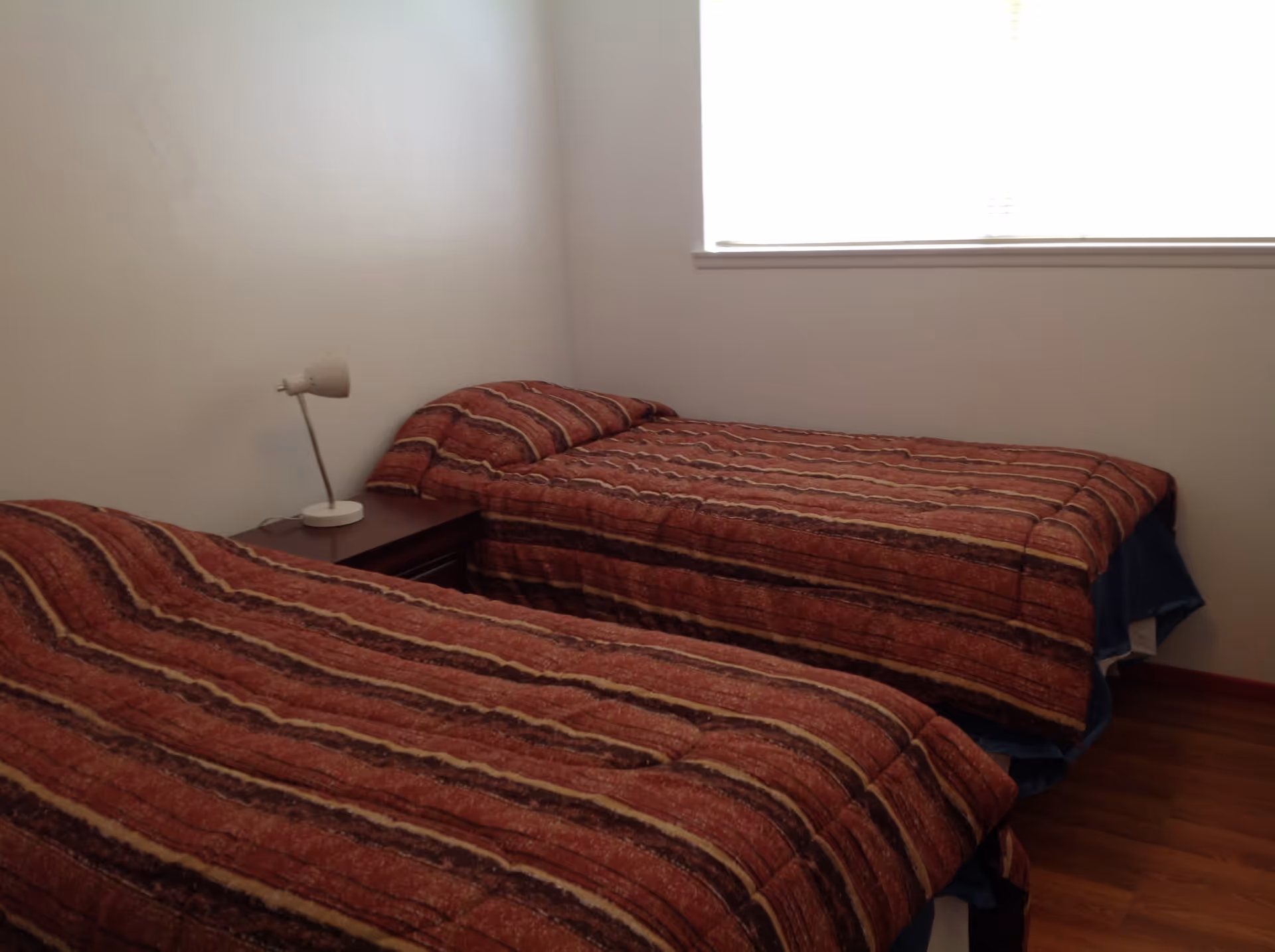 A simple bedroom with two single beds covered in matching red and brown striped bedspreads. A small wooden nightstand with a white lamp is positioned between the beds. There is a window with blinds on the wall behind one of the beds, and the floor is wooden.