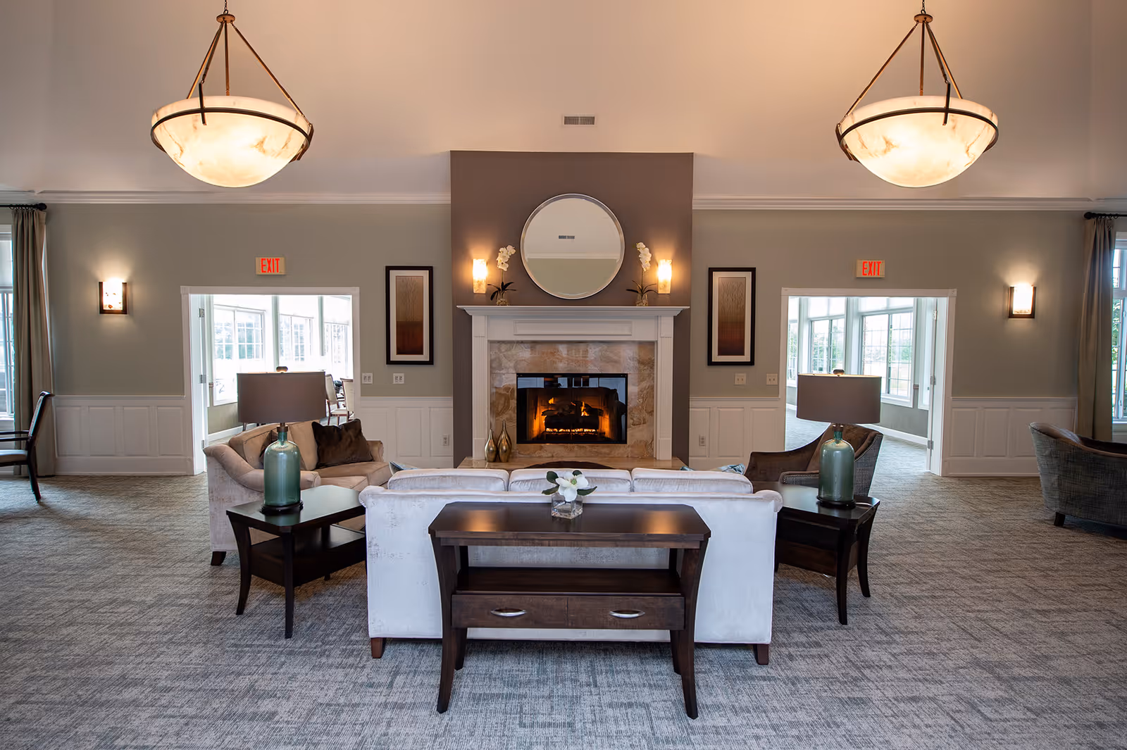 A spacious and well-lit living room area with a central fireplace surrounded by a beige sofa and armchairs. Two side tables with lamps flank the sofa, and a round mirror hangs above the fireplace. The room features large windows, neutral-colored walls, carpeted flooring, and two large ceiling light fixtures.