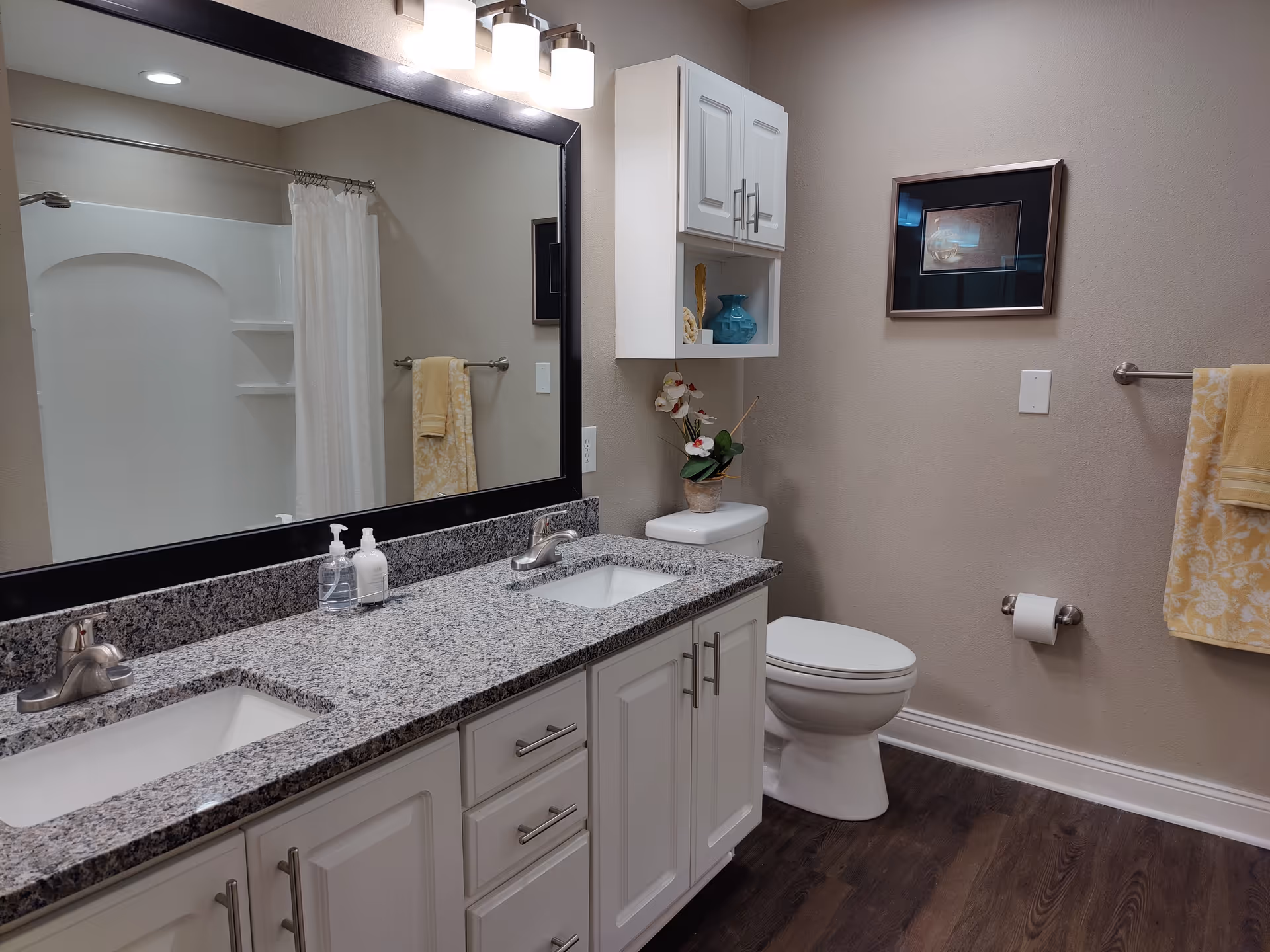 A clean and modern bathroom with a double sink vanity featuring a granite countertop and white cabinets. Above the sinks is a large mirror with three light fixtures. To the right, there is a toilet with a small cabinet mounted above it, decorated with a blue vase and flowers. A towel rack with yellow patterned towels and a toilet paper holder are mounted on the beige wall. The floor is dark wood, and a shower with a white curtain is visible in the mirror reflection.