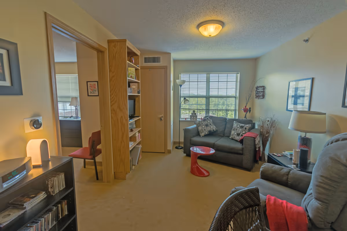 A cozy living room with a gray couch adorned with patterned pillows and a red throw blanket, a red modern side table, a gray recliner chair with a red blanket, a black side table with a lamp and decorative items, a tall floor lamp near a large window with blinds, and a wooden bookshelf with various items. The room has beige walls and carpet, and an open doorway leads to another room with a red chair and a window with blinds.