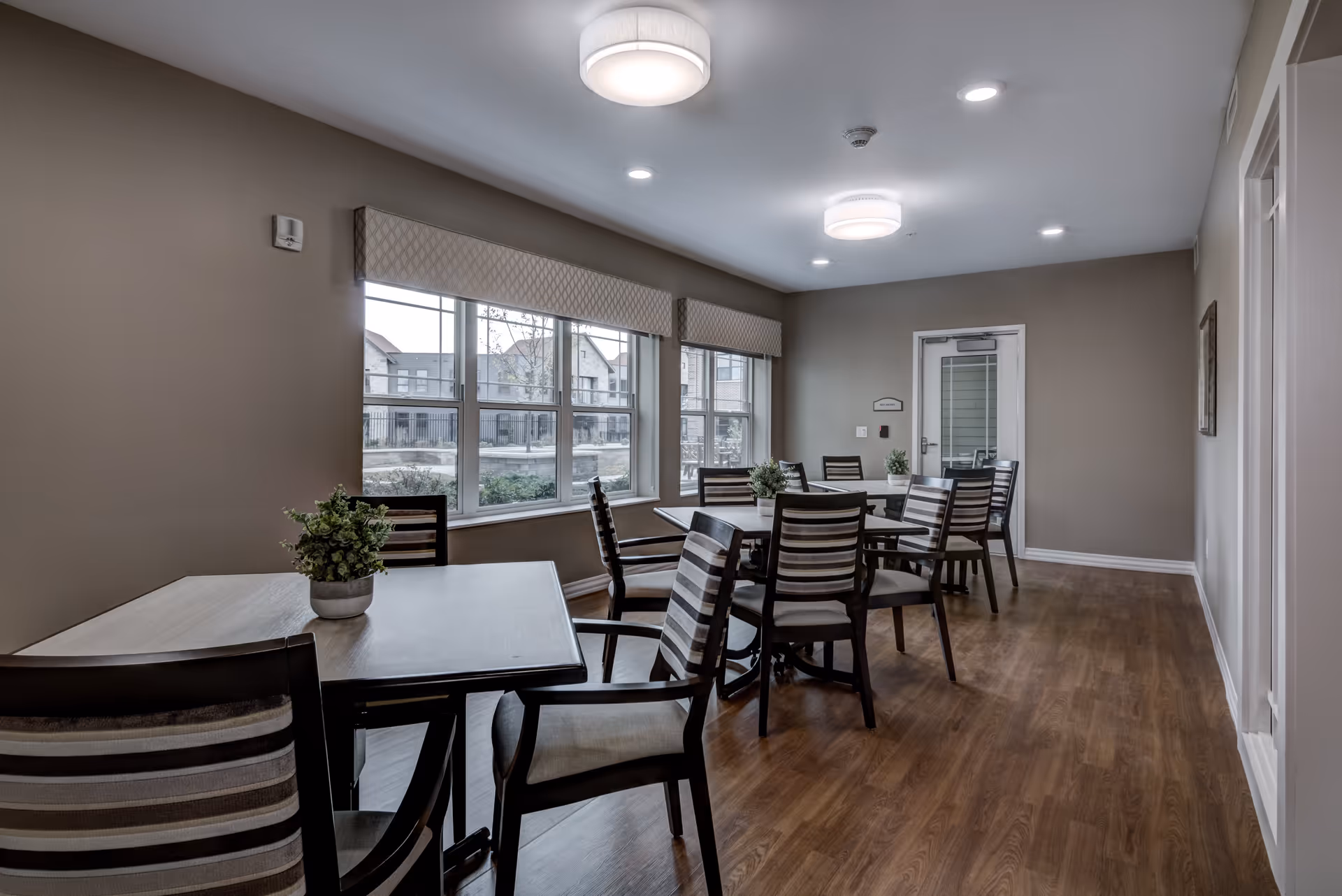 A dining room in Azura Assisted Living and Memory Care of Fox Point with several wooden tables and striped cushioned chairs arranged neatly. Each table has a small potted plant centerpiece. Large windows with patterned valances allow natural light to brighten the room, and the floor is covered with wood-style flooring. The walls are painted beige, and ceiling lights provide additional illumination.