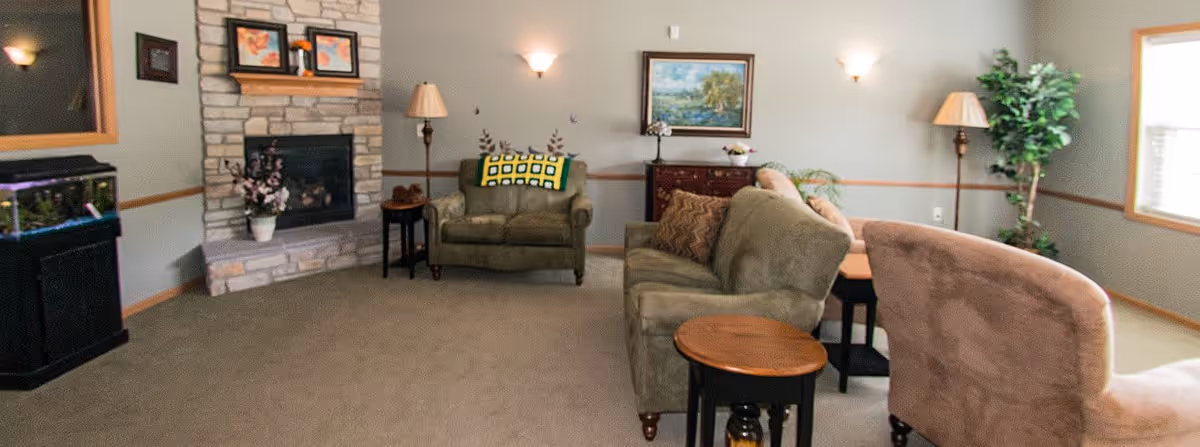 A cozy living room with a stone fireplace, a green armchair with a yellow and green blanket, a green sofa with brown cushions, a beige armchair, wooden side tables, a floor lamp, a potted plant, a fish tank, and a painting on the wall.