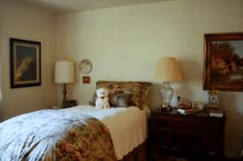 A cozy bedroom with a single bed covered in floral bedding and a white blanket. A stuffed dog toy is placed on the bed. There are two bedside tables, each with a lamp, and framed artwork hanging on the walls.