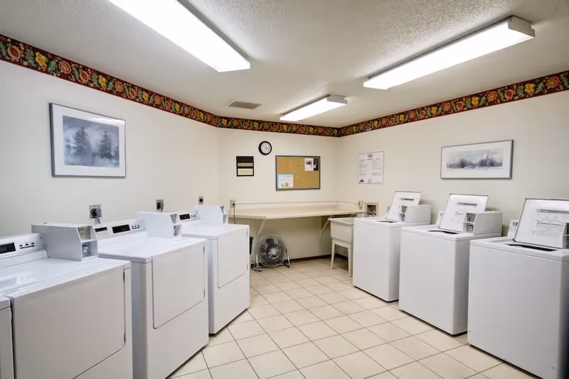 Laundry room with six white washing machines and dryers arranged along two walls. The room has tiled flooring, white walls with a decorative floral border near the ceiling, fluorescent ceiling lights, a wall clock, bulletin boards, and framed pictures on the walls. There is also a small fan and a folding table in the corner.