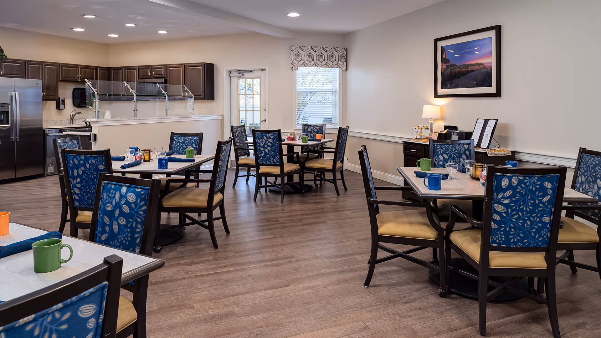A dining room in a senior living facility with several tables and chairs. Each table is set with colorful mugs, glasses, and napkins. The room features wood flooring, a kitchen area with dark cabinets and stainless steel appliances, and a window with a patterned valance. A framed picture and a lamp are visible on a sideboard against the wall.