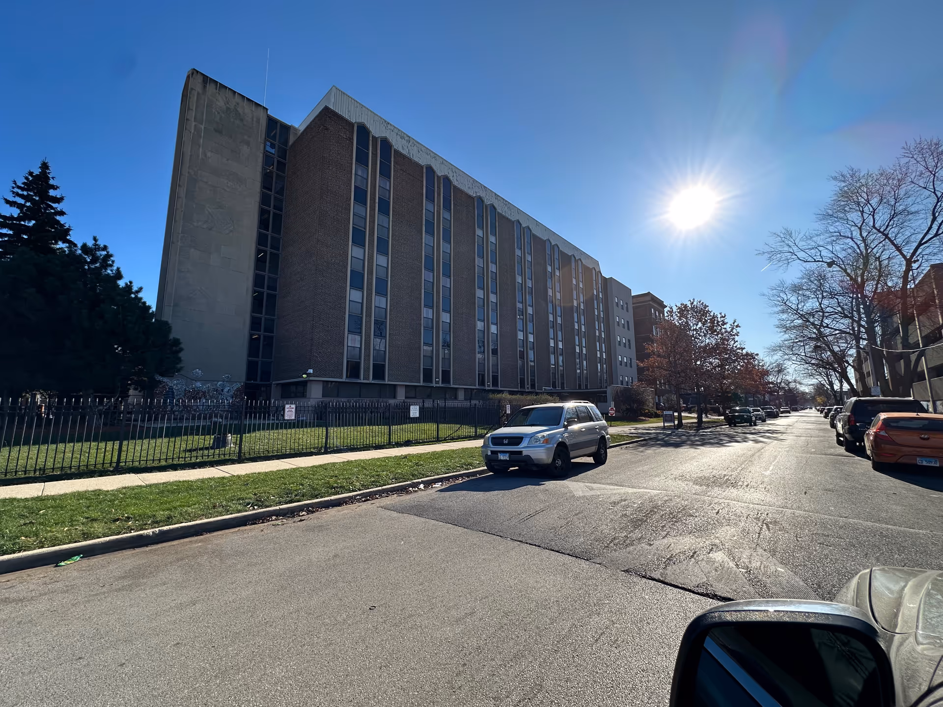 Exterior view of a multi-story brick building under a clear blue sky with the sun shining brightly. The building is surrounded by a black metal fence and there are several cars parked along the street in front of it. Trees with sparse leaves line the sidewalk.