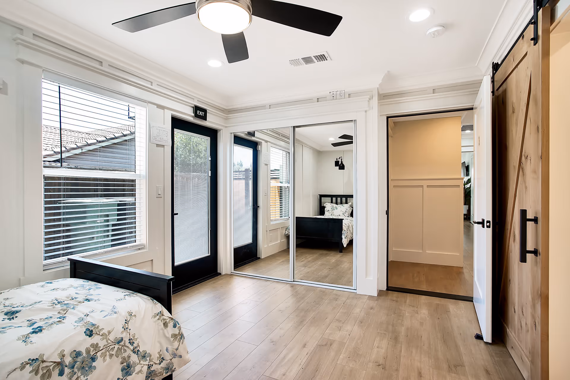 A bright bedroom with light wood flooring, a black bed frame with floral bedding, a ceiling fan with light, large windows with blinds, a black door with an exit sign above it, and a mirrored closet door reflecting the bed and part of the room.