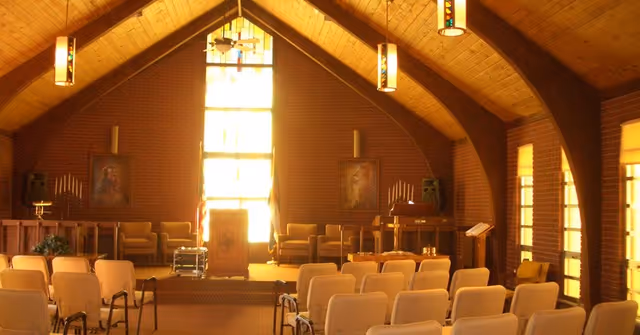 Interior of a chapel or worship room with wooden vaulted ceiling, stained glass window at the front, rows of beige chairs facing a wooden podium, and religious paintings on the brick walls.