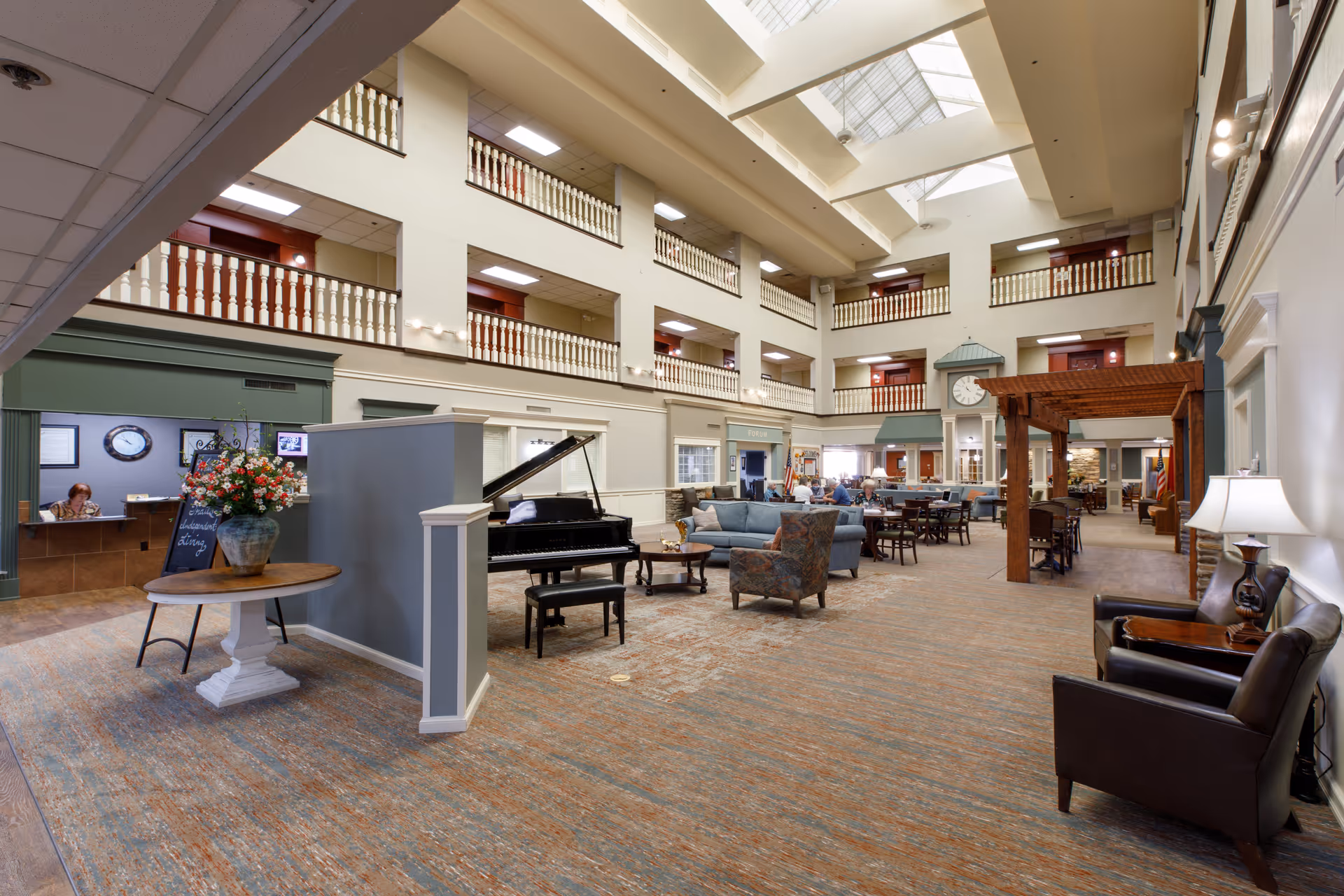 Spacious and well-lit common area in a senior living facility with high ceilings and skylights. The room features a grand piano, comfortable seating including sofas and armchairs, tables and chairs for socializing, a reception desk with a staff member, and decorative elements such as a large flower arrangement and a clock tower structure. The area is surrounded by multiple floors with balconies overlooking the space.