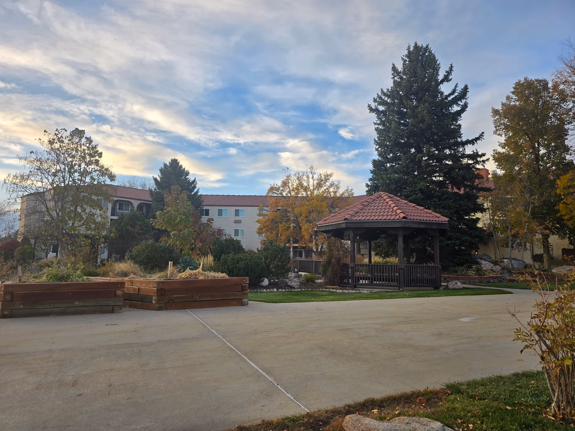 Outdoor courtyard with a gazebo, raised planter beds, trees, and a multi-story building under a cloudy sky.