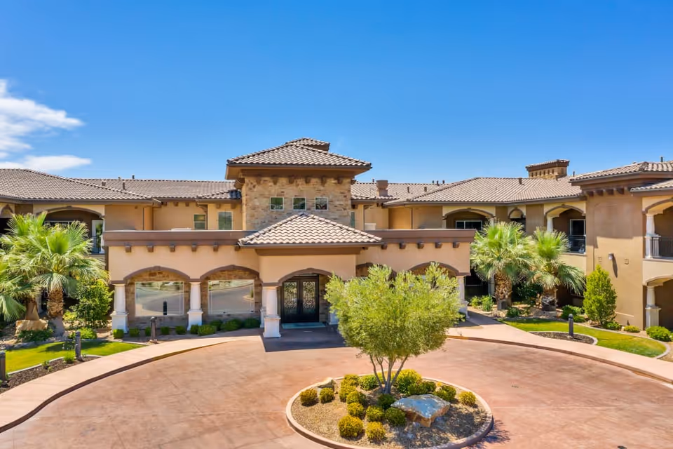 Front exterior view of The Retreat at Sunbrook facility featuring a circular driveway with a landscaped center island, palm trees, and a building with a tiled roof and arched entryways under a clear blue sky.