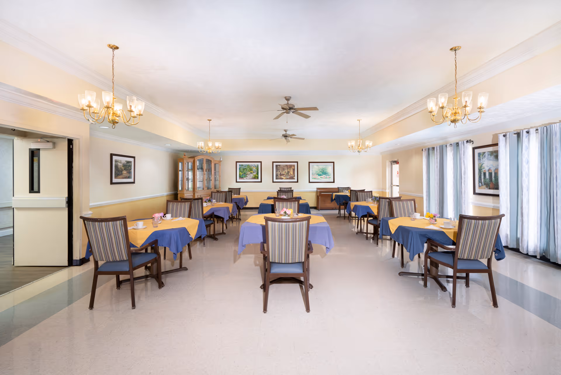 A bright dining room with several tables covered with blue and yellow tablecloths, each set with cups and small flower arrangements. The room has striped cushioned chairs, chandeliers hanging from the ceiling, ceiling fans, framed artwork on the walls, and large windows with light curtains allowing natural light to enter.