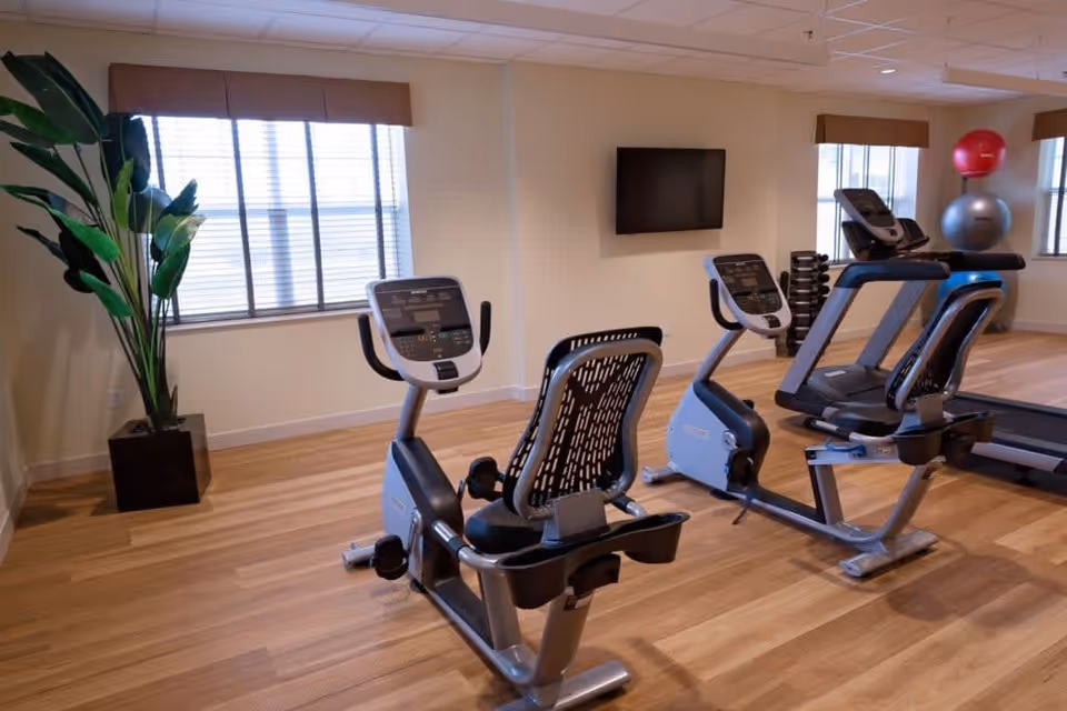 Bright exercise room with recumbent bikes, treadmills, exercise balls, and a wall-mounted TV.