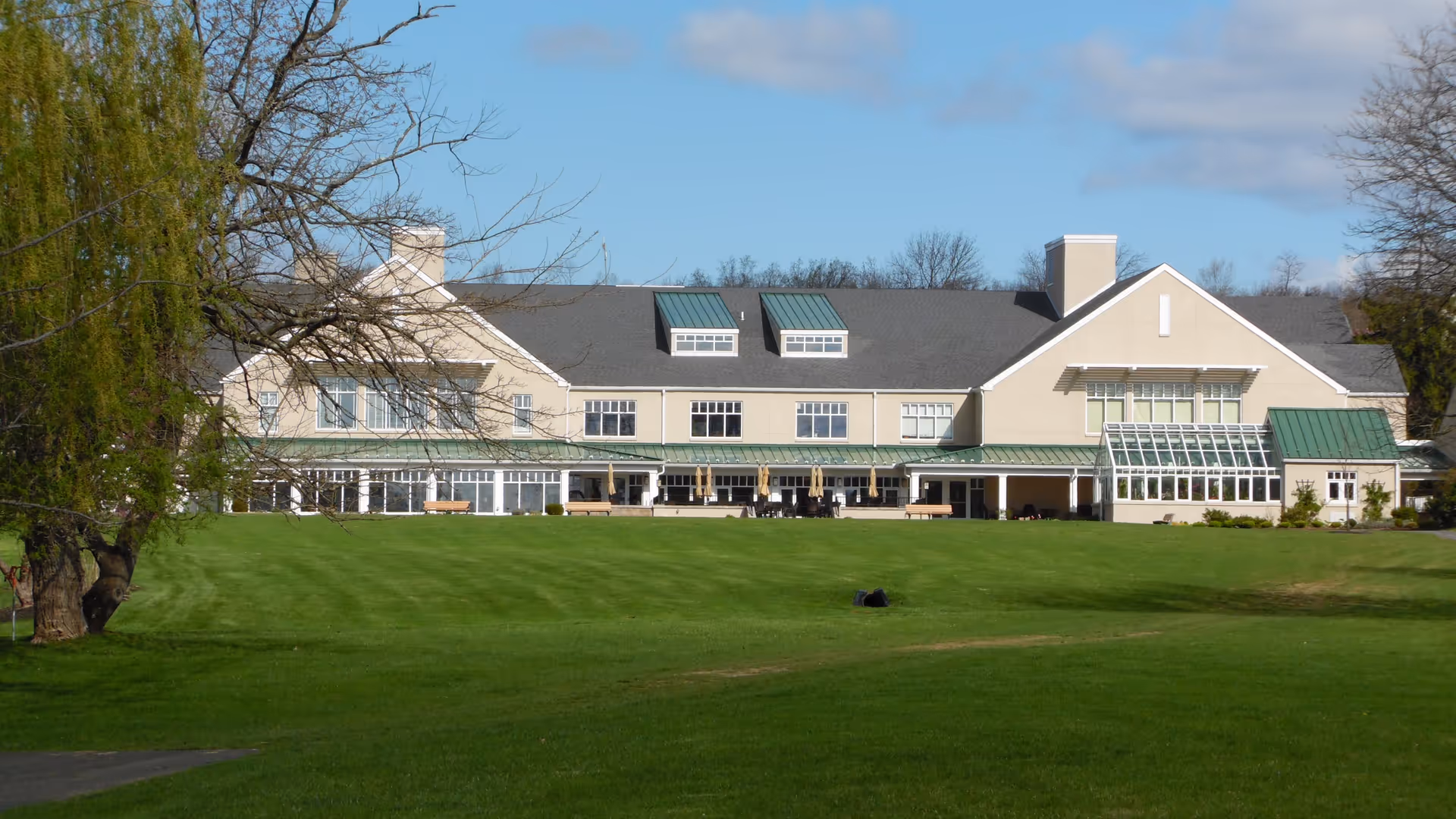 Exterior view of a large beige building with green roofs and multiple windows, surrounded by a well-maintained green lawn and trees under a partly cloudy sky.